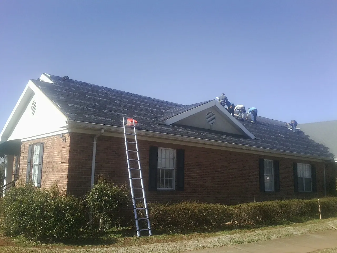 Crew tearing shingles off a brick ranch roof with a ladder set — Metal Roof Repair work by our crew in Exline