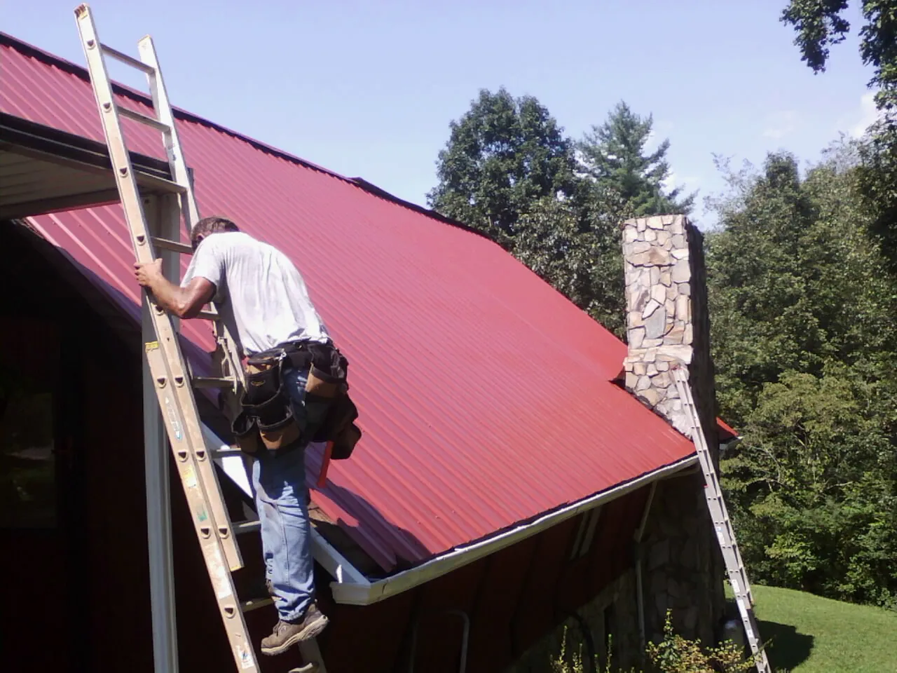 Roofer on a ladder beside a red metal roof and stone chimney — Soffit & Fascia Repair by Roofing Brothers in Exline
